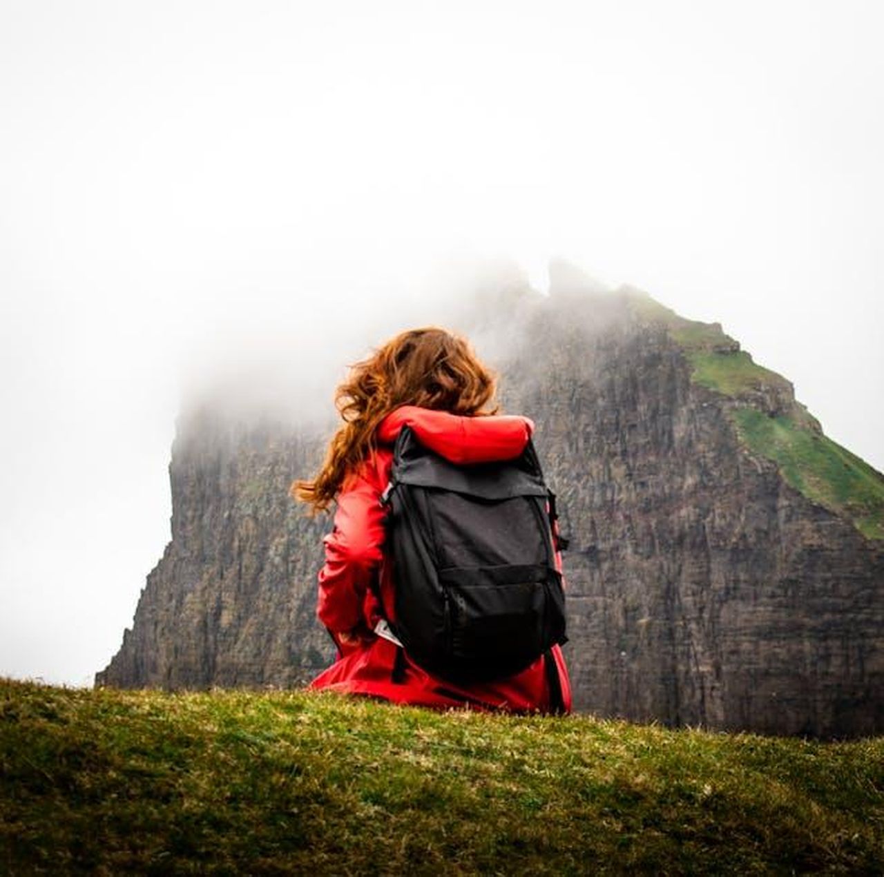 3_Georgia_parte_per_un_viaggio_da_sola_i7qSybCBt5 - Confidenze24 Source: Tomáš Malík - pexels - A woman sits with a backpack in front of fog-covered cliffs in the Faroe Islands, perfect for adventure travel themes.