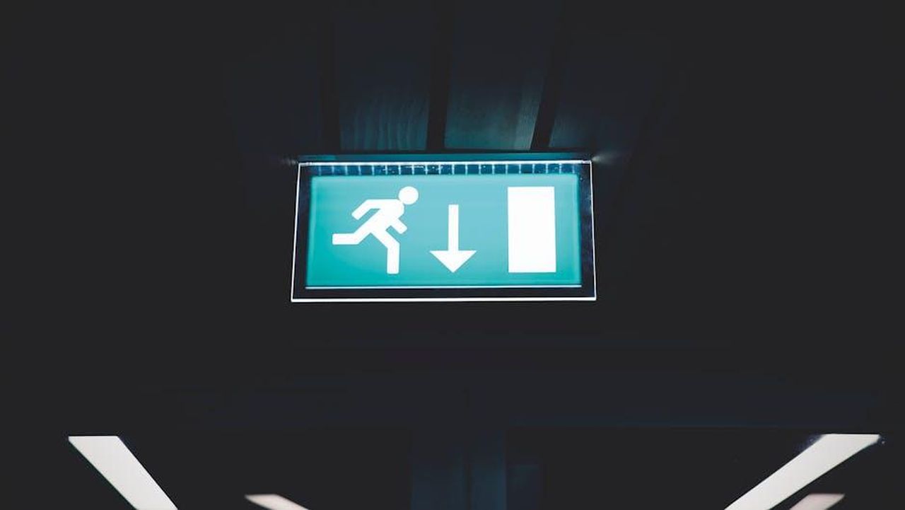 4_Tracy_compiacente_vj7Y0u5GDR - Confidenze24 Source: Matthias Zomer - pexels - Brightly lit emergency exit sign with arrow indicating direction, set against a dark backdrop.