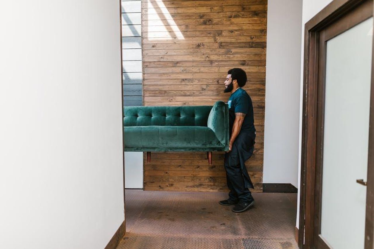 6_Georgia_si_trasferisce_dalla_nuova_casa_gkCBBKsxid - Confidenze24 Source: RDNE Stock project - pexels - A mover carries a green sofa indoors, showcasing modern interior design and relocation services.