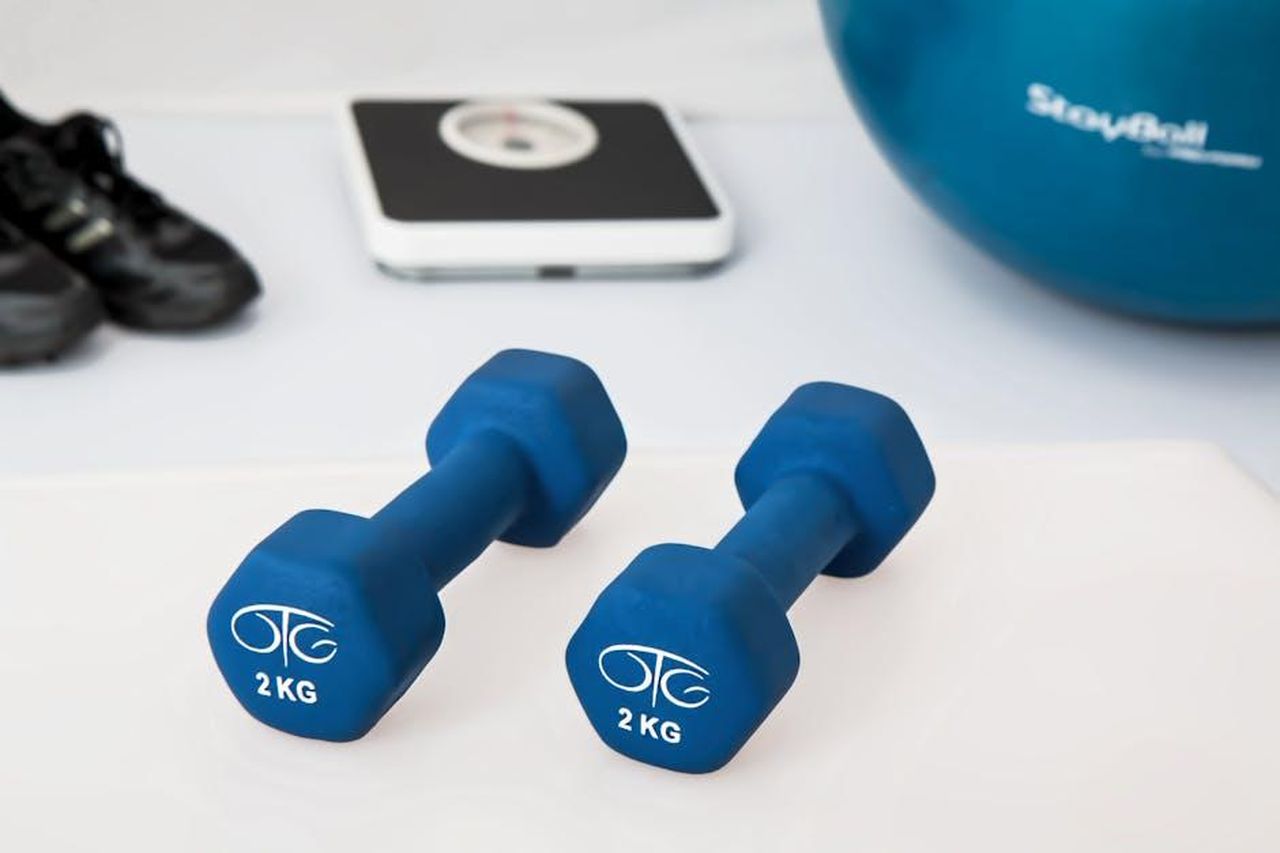 6_Riabilitazione_X7JA7SC0tA - Confidenze24 Source: Pixabay - pexels - Blue dumbbells placed on a white surface with fitness equipment in the background.