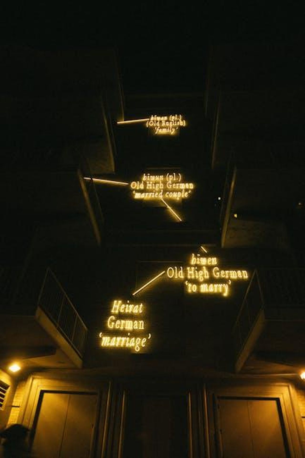 6__di_origine_greca_3NpzbgywZs - Confidenze24 Source: Athena Sandrini - pexels - Stunning nighttime display of illuminated neon signs exploring German etymology on a building.