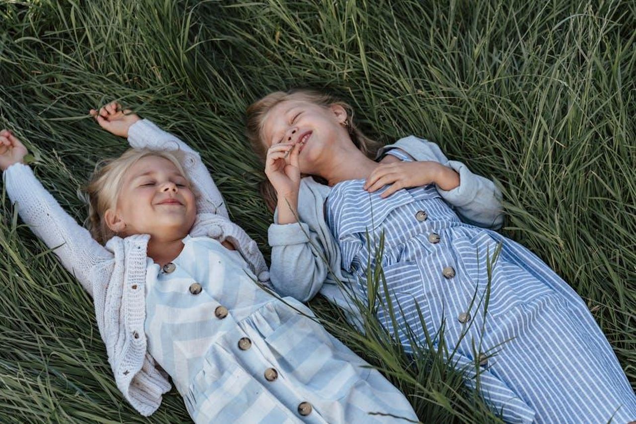 Abbigliamento_estivo_e_vestiti_o80cHx5TMg - Confidenze24 Source: olia danilevich - pexels - Two young girls happily lying on grass, enjoying a playful moment together.
