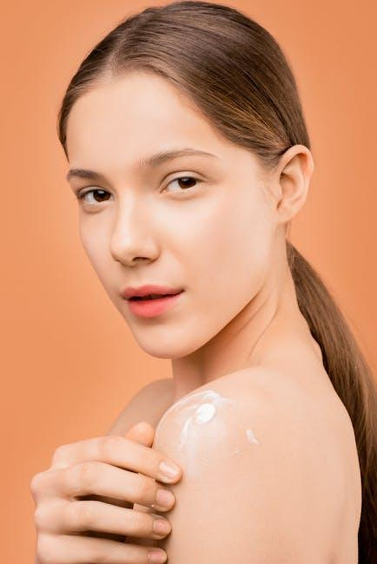 Adatto_a_tutti_i_tipi_di_pelle_HDk2XPIzmj - Confidenze24 Source: Shiny Diamond - pexels - Young woman applies moisturizer on shoulder, set against a peach background, highlighting smooth skin care.