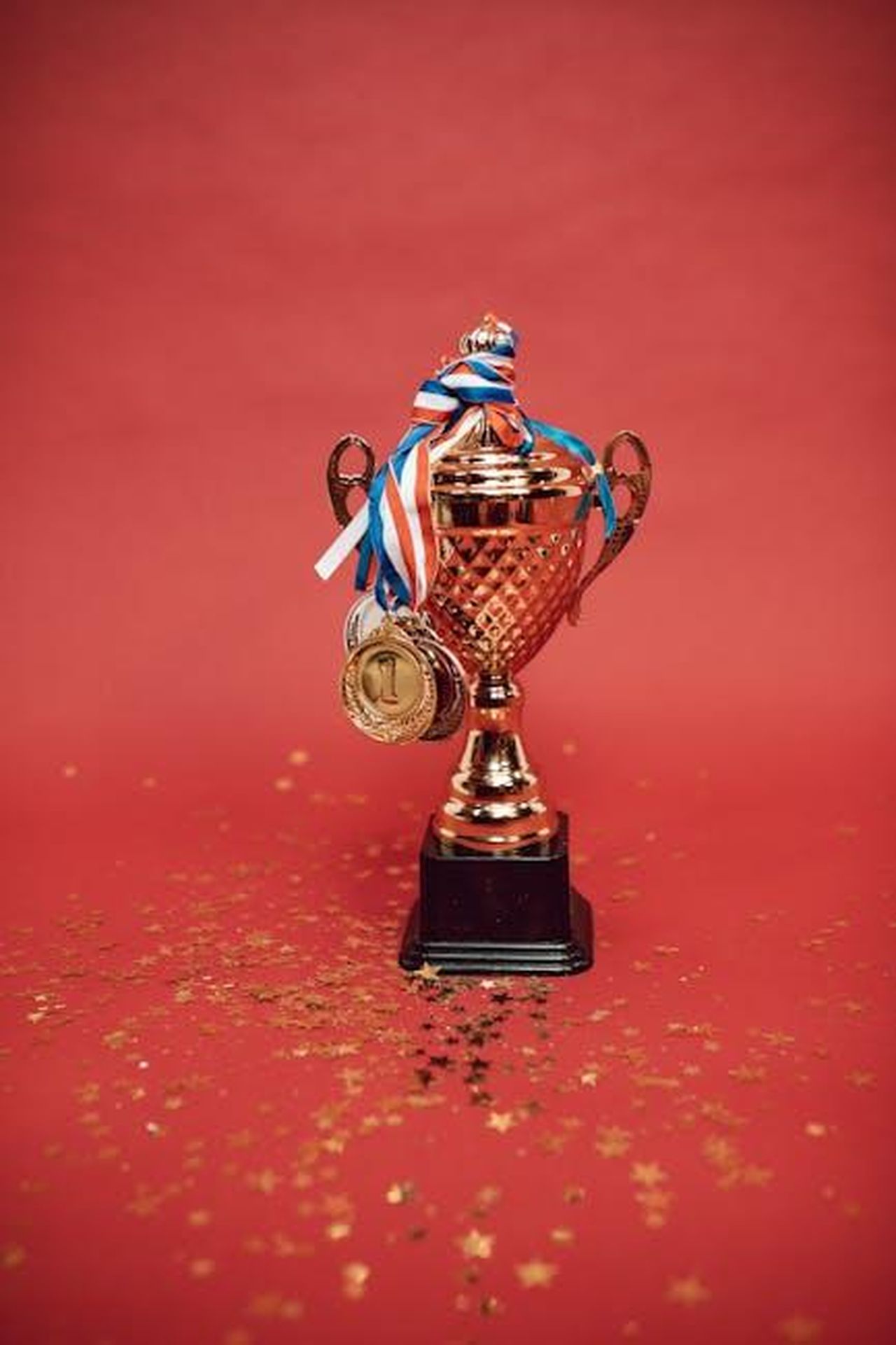 Ben_rivela_il_premio_fantastico_del_giocatore_xzYo0F73TQ - Confidenze24 Source: RDNE Stock project - pexels - Golden trophy adorned with medals on a red backdrop surrounded by scattered confetti.