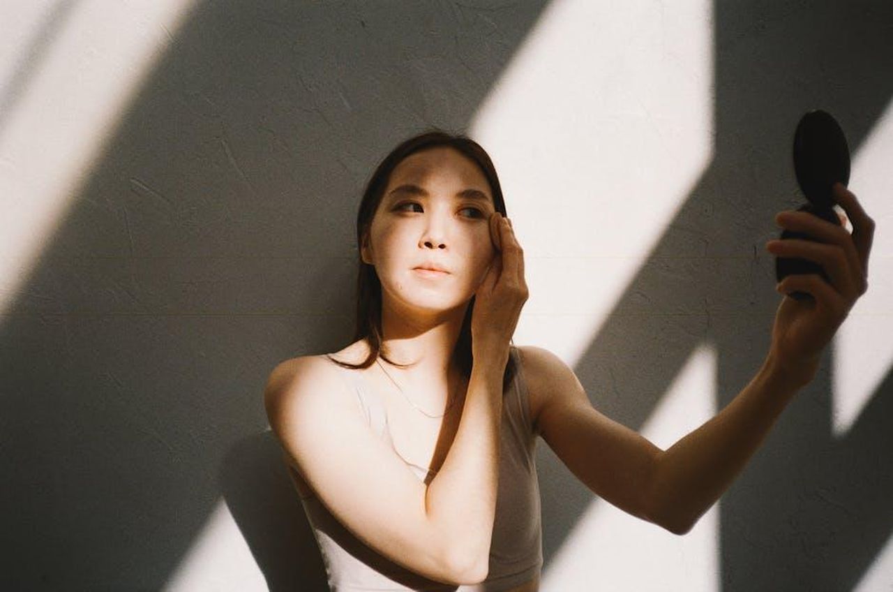 Caratteristiche_uniche_del_prodotto_de1JQzpJPw - Confidenze24 Source: Polina Tankilevitch - pexels - A young Asian woman applies makeup with natural sunlight highlighting her face indoors.