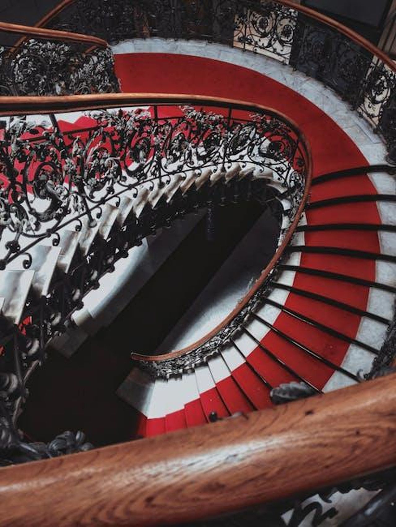 Celebrit_sul_Red_Carpet_FbIV1uiEC4 - Confidenze24 Source: Matheus Viana - pexels - A stunning spiral staircase with intricate wrought iron railings and a vibrant red carpet, viewed from above.