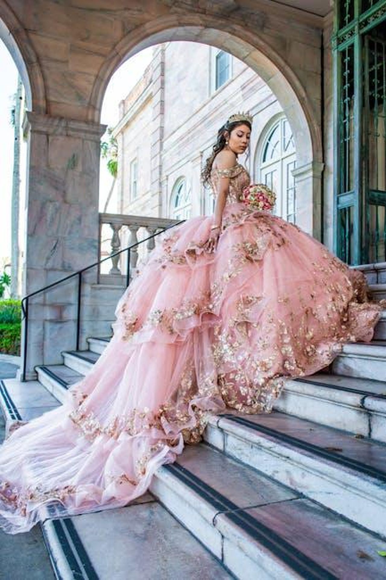 Chi__LuciaSofia_Ponti_e_il_suo_debutto_pubblico_sdziXP9s2A - Confidenze24 Source: Johana A. - pexels - A young woman in an elaborate pink quinceañera dress poses elegantly on castle stairs, showcasing floral details.