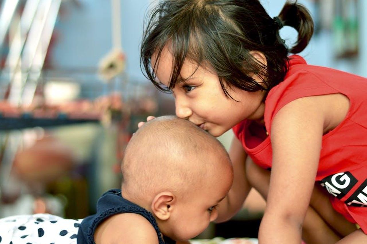 Chi__la_nuova_fidanzata_di_James_P5LzHFnXif - Confidenze24 Source: Diwakar Jangir - pexels - A touching moment of sisterhood as a young girl gently kisses her baby brother's head indoors.