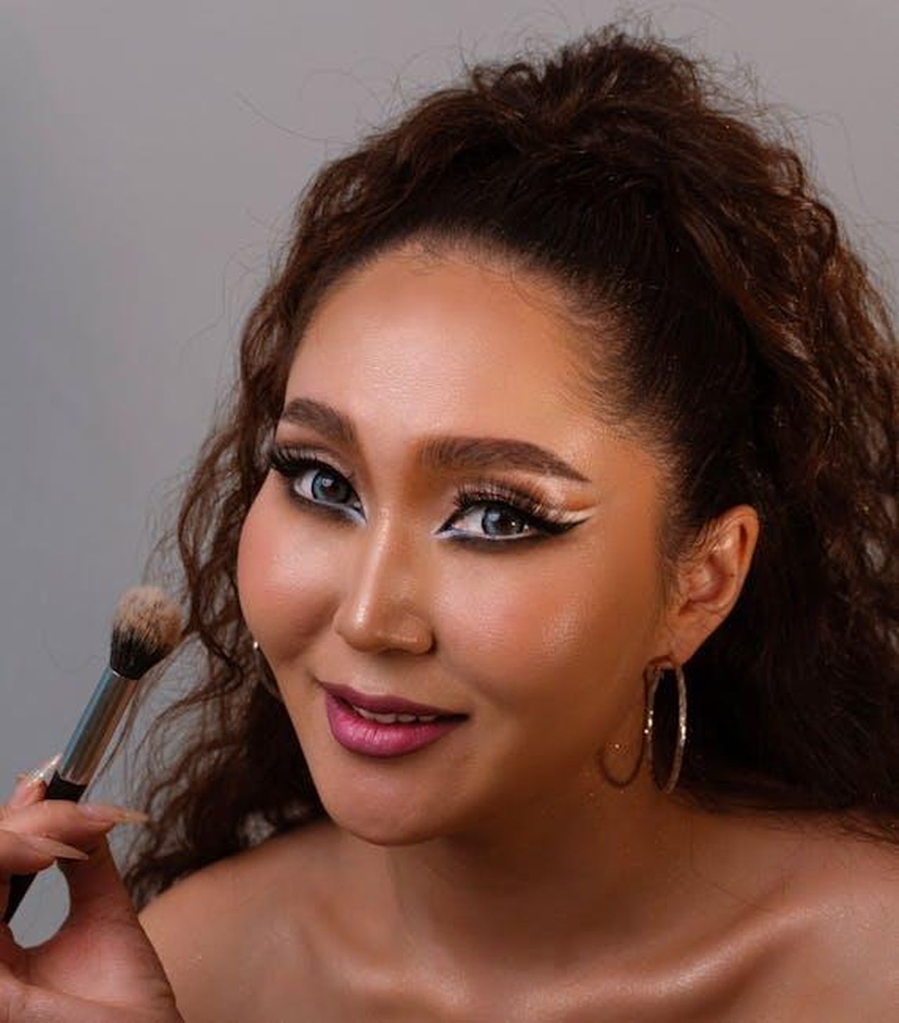 Come_Applicare_il_Fondotinta_Correttore_jFna7zBOlU - Confidenze24 Source: BYB BYB - pexels - A woman smiles confidently while holding a makeup brush, showcasing bold makeup.