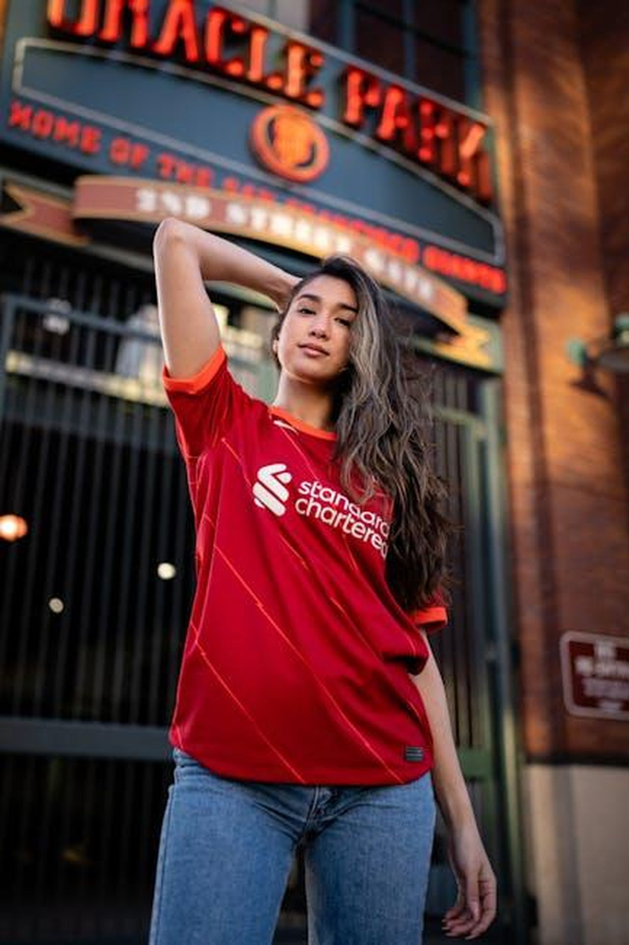 Coronation_Street_i_fan_prevedono_una_trama_di_aggressione_sessuale_per_Mick_Michaelis_oyJzINils5 - Confidenze24 Source: Marcus Queiroga Silva - pexels - Confident woman posing in a Liverpool FC jersey outside a stadium entrance.