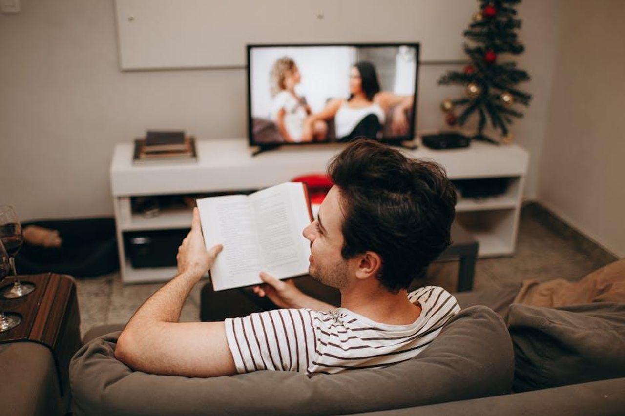 Cosa_aspettarsi_dalla_trama_della_settima_stagione_jO0kcOWGkO - Confidenze24 Source: Helena Lopes - pexels - A man sits on a sofa reading a book in a cozy living room with a TV on.