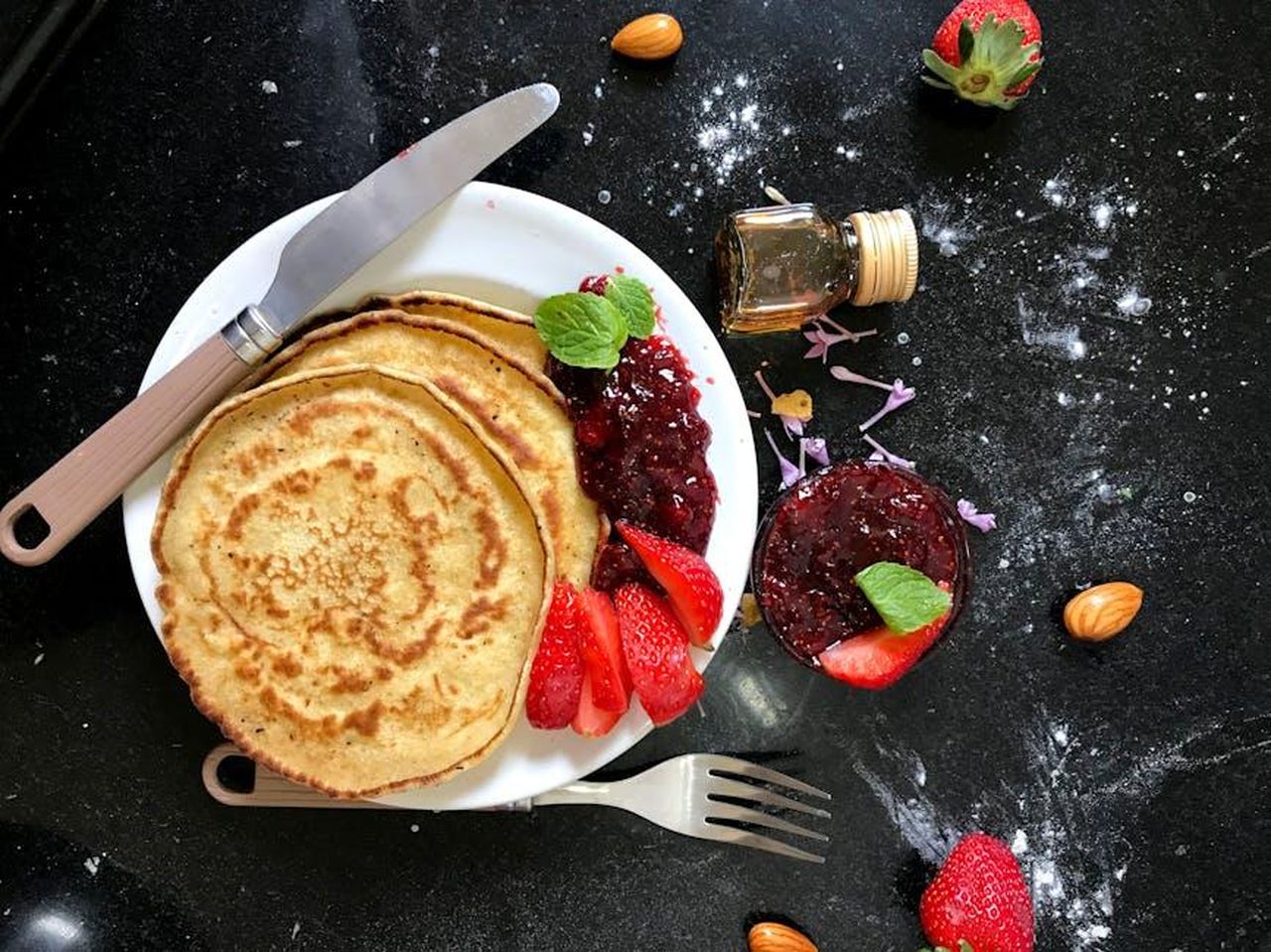 Cosa_mangiano_i_Sussex_a_colazione_pCkQc2PTT6 - Confidenze24 Source: Rama Khandkar - pexels - Top view of pancakes with strawberry jam, fresh strawberries, almonds, and honey on a dark kitchen counter.
