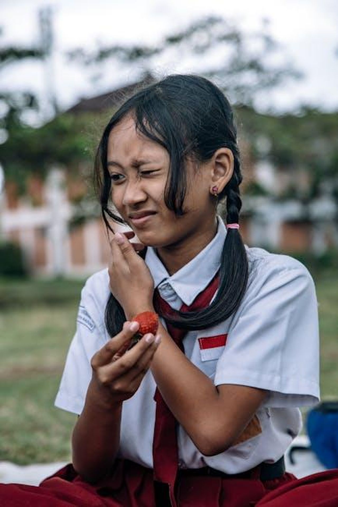 Critiche_alla_girl_squad_di_Taylor_4M6CsSr2IY - Confidenze24 Source: ROMAN ODINTSOV - pexels - A young girl in a school uniform holding a fruit in an outdoor setting, displaying a playful expression.