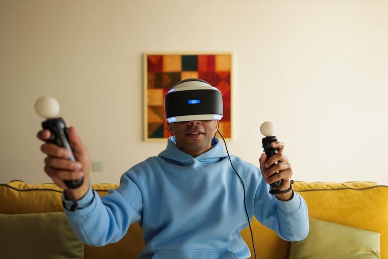 Critiche_di_Sharon_ai_programmi_di_Simon_pgUo1WjpOM - Confidenze24 Source: VAZHNIK - pexels - Adult man wearing VR headset and holding controllers, fully immersed in virtual reality gaming in a cozy living room.