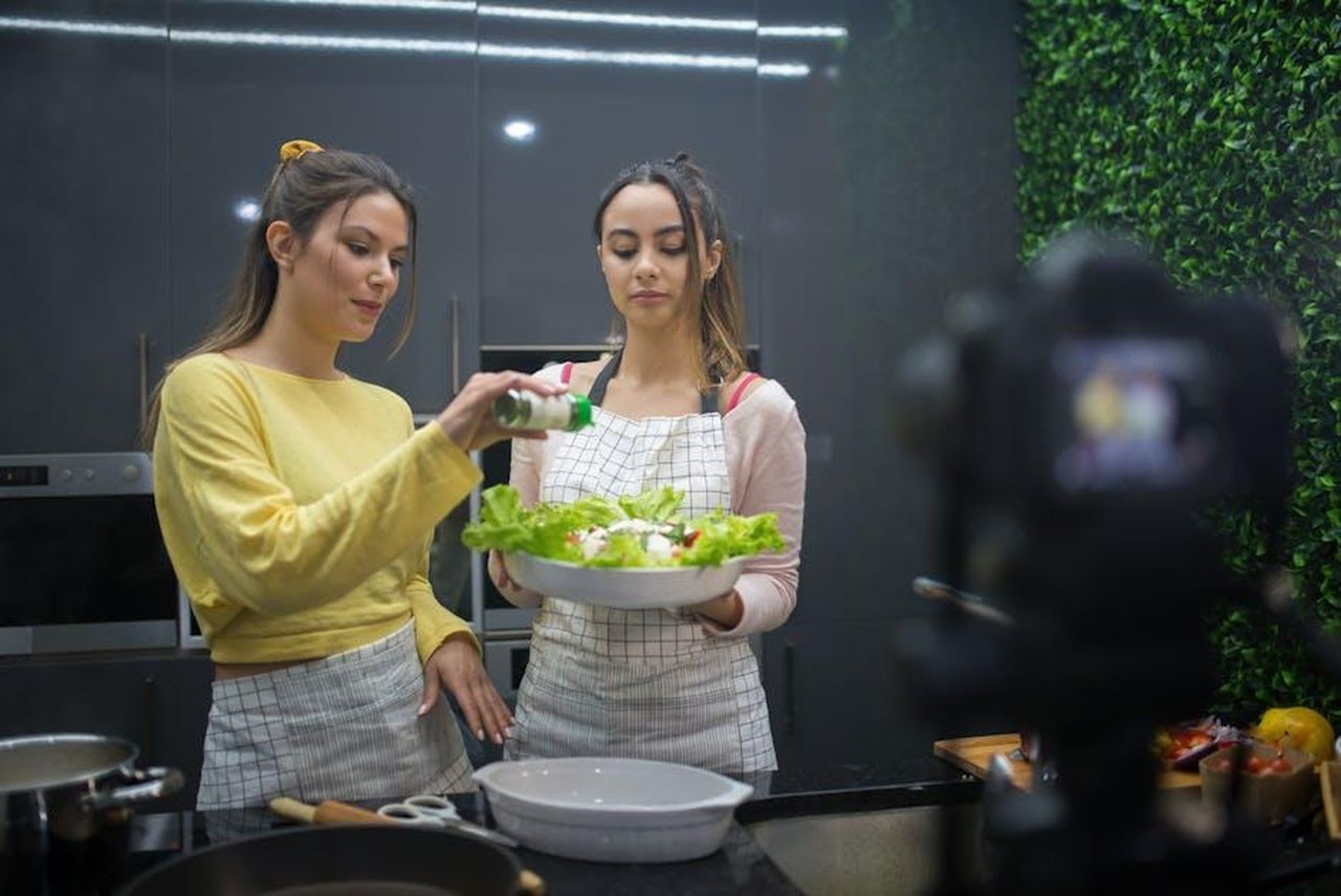 Critiche_internazionali_e_confronti_con_altri_show_CZ4qdLYu0J - Confidenze24 Source: Kampus Production - pexels - Two women preparing a salad while recording a cooking video in a stylish kitchen setting.