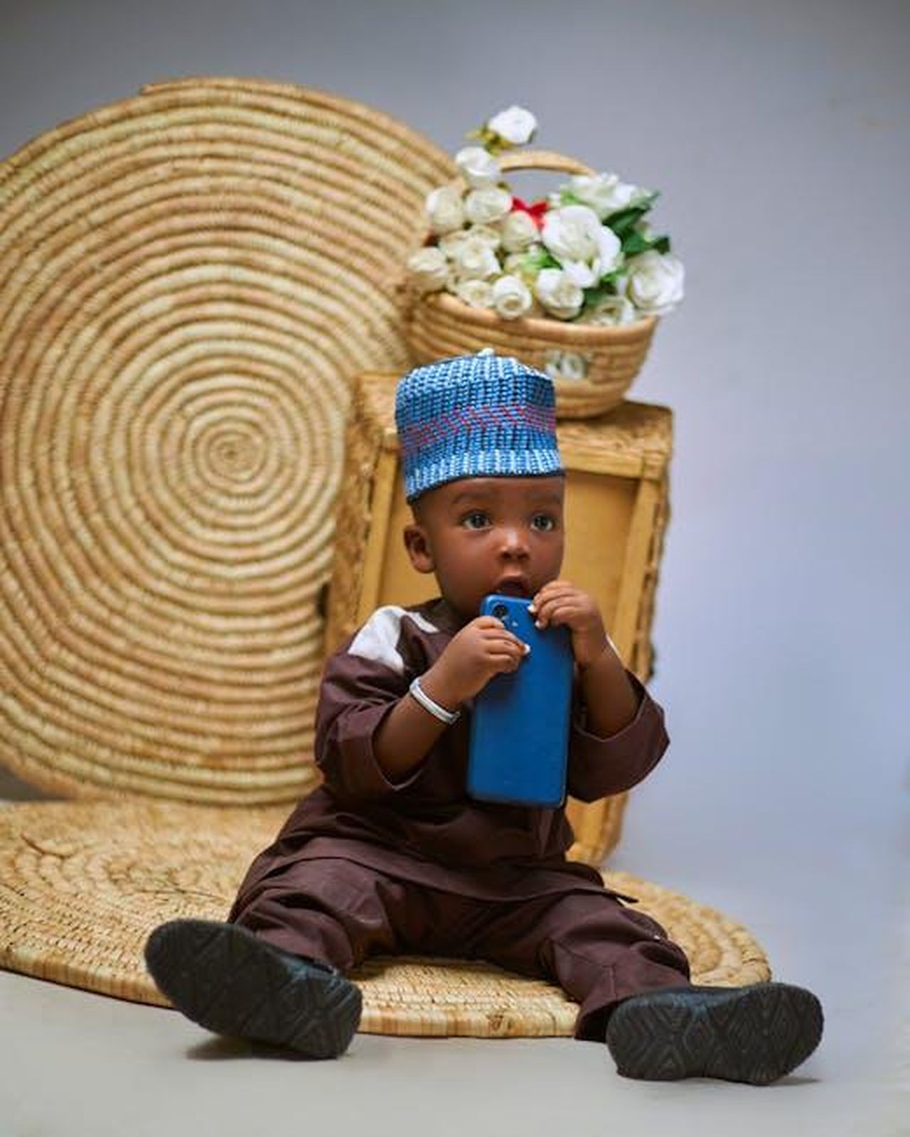Critiche_per_la_foto_di_Harry_e_Lilibet_4dWTm9fMam - Confidenze24 Source: Boko Shots - pexels - Cute baby in traditional attire holding a phone, sitting with roses.