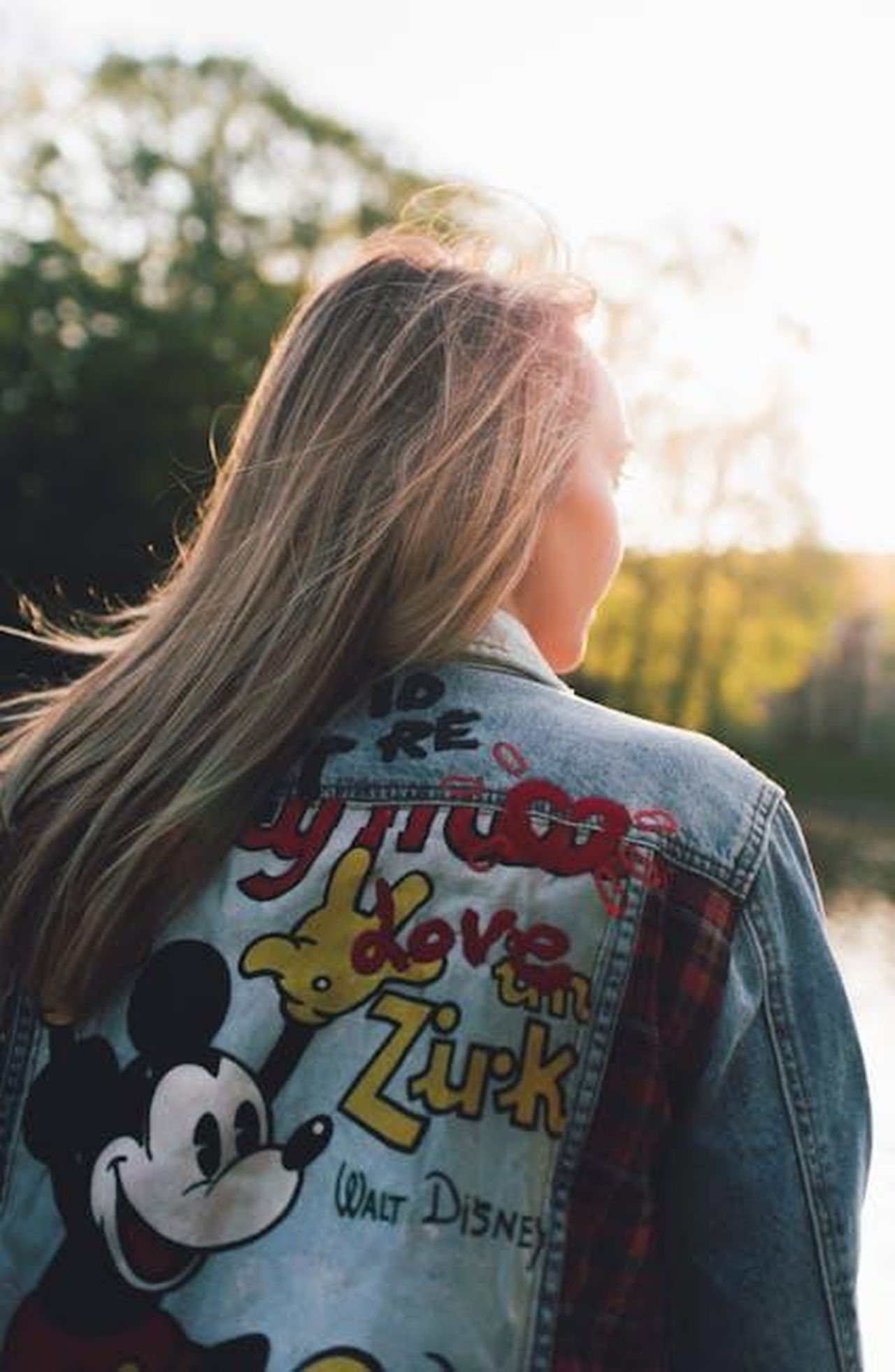 Design_e_caratteristiche_della_collezione_LlsNrs6Sbs - Confidenze24 Source: Iurii Laimin - pexels - A woman with long blonde hair wearing a denim jacket featuring a colorful Mickey Mouse design in an outdoor setting.