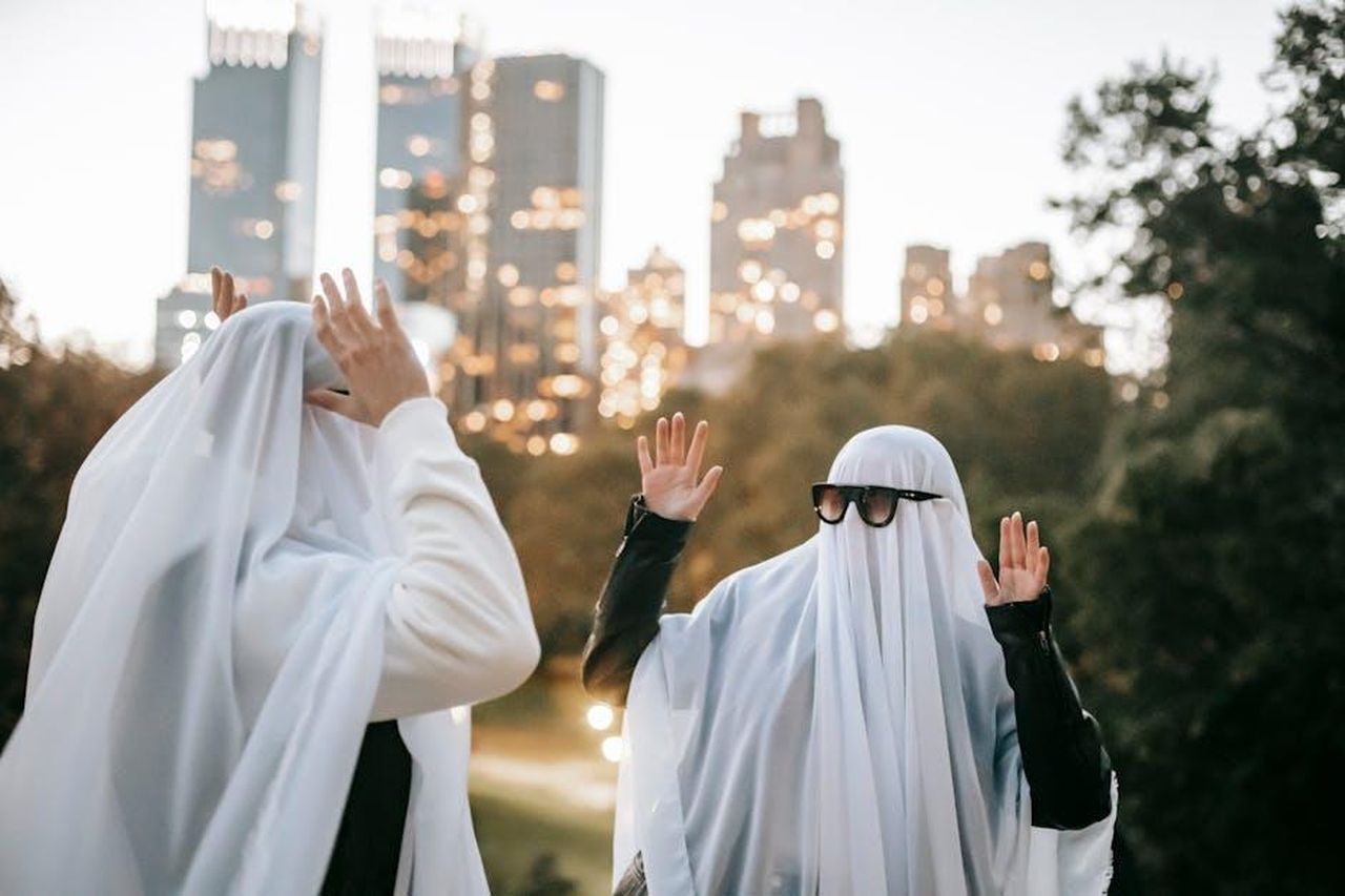 Dettagli_sui_nuovi_personaggi_BuzxJUkyCC - Confidenze24 Source: Katerina Holmes - pexels - Funny unrecognizable couple with raised arms wearing ghost costumes and sunglasses looking at each other while standing in park with glowing skyscrapers on blurred background during Halloween