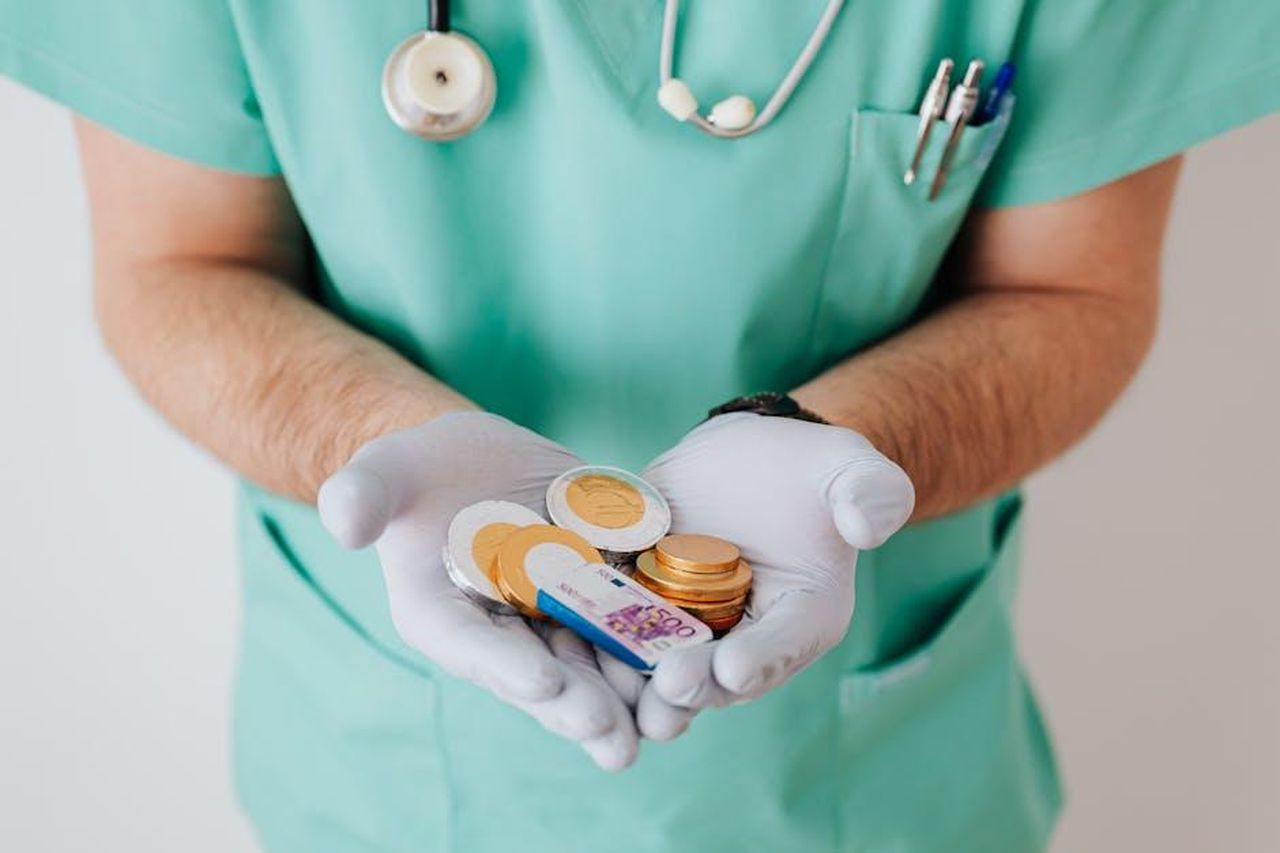 Difficile_giustificare_il_costo_8JmCQyrOIe - Confidenze24 Source: Photo By: Kaboompics.com - pexels - Unrecognizable medical worker demonstrating money wearing medicine form standing near white wall in hospital while working in day of salary