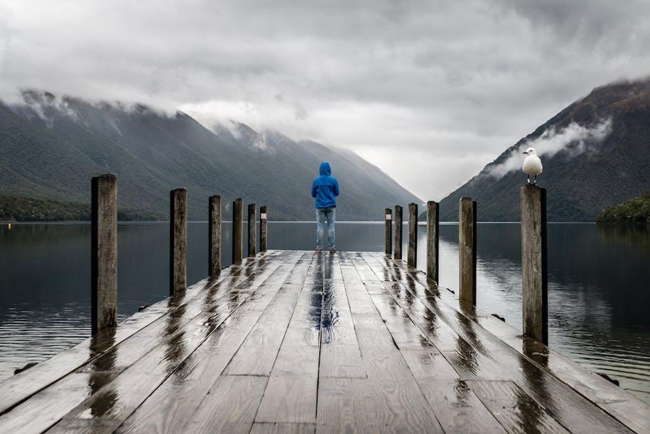 Dimostra_limpatto_sul_loro_umore_uQODojO5ov - Confidenze24 Source: Gabriela Palai - pexels - Person in blue jacket stands on a rainy dock overlooking serene lake and mountains.