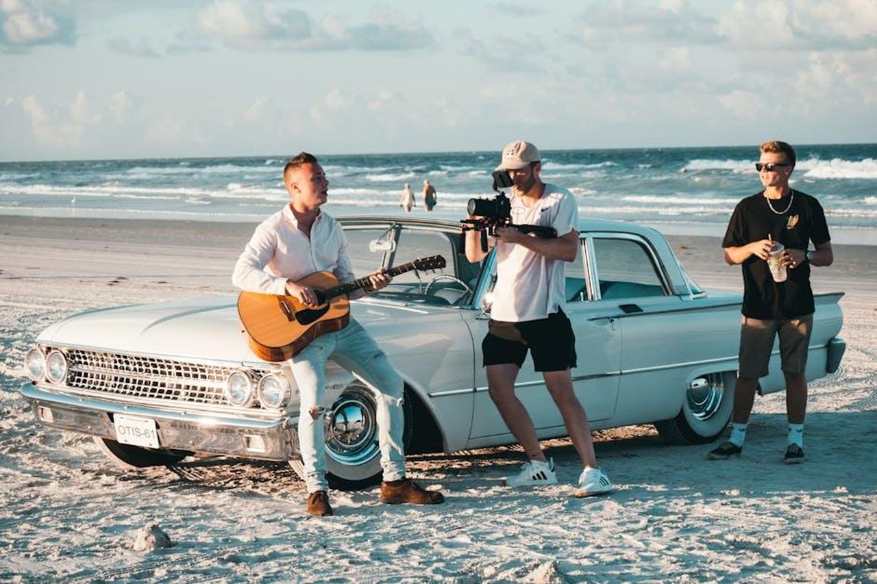 Dr_G_Si_Sente_Minacciato_E_Pubblica_Una_Canzone_Ss2jzQxGsh - Confidenze24 Source: Kyle Loftus - pexels - A musician plays guitar on a beach as a filmmaker records with a camera beside a classic car.