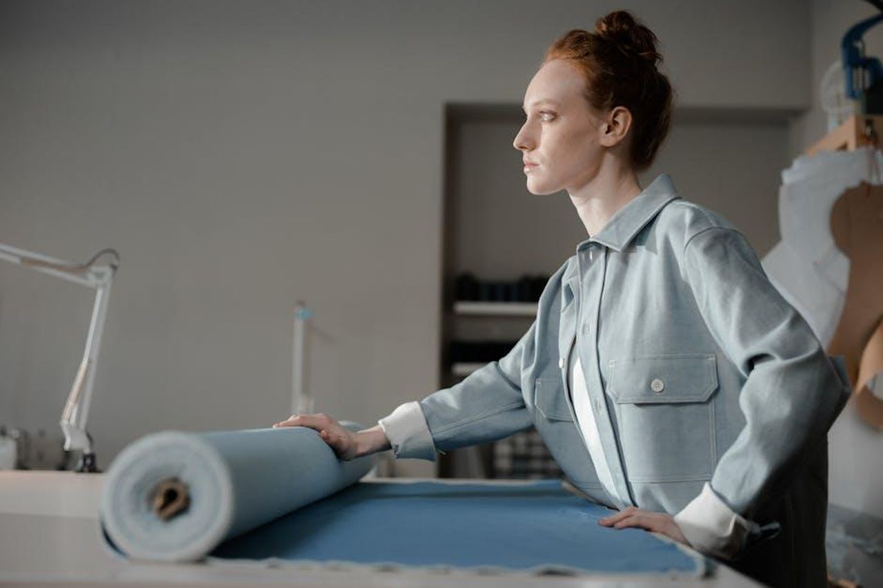 Evoluzione_dello_stile_di_Shiloh_cXm4lj4o5j - Confidenze24 Source: Ron Lach - pexels - A female fashion designer positioned side view, holding a roll of blue fabric in a studio setting, showcasing creativity.