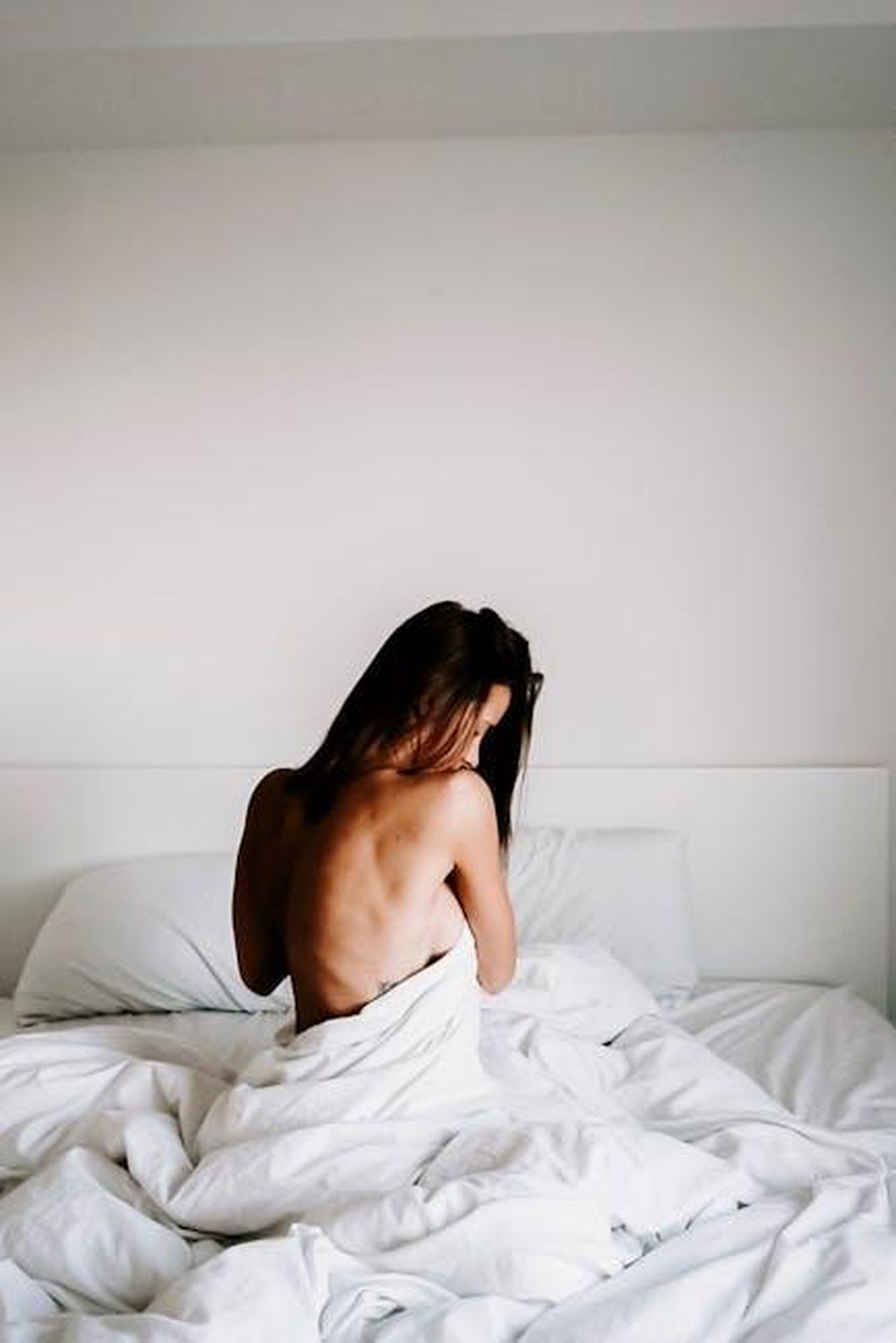 Fuga_di_foto_nuda_4OYCWMXqQn - Confidenze24 Source: Julia Kuzenkov - pexels - A serene, minimalist portrait of a woman sitting on a bed, enveloped in soft white linens, in a tranquil bedroom setting.