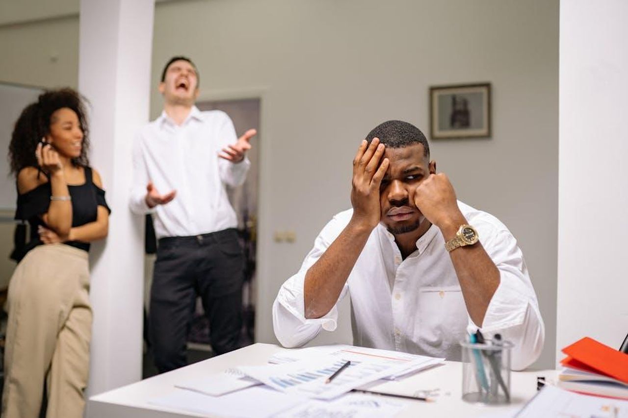 Gary_Linekar_outing_la_relazione_iBV53YGPlV - Confidenze24 Source: Yan Krukau - pexels - A man at a desk looks frustrated while two coworkers laugh. Office workplace conflict concept.