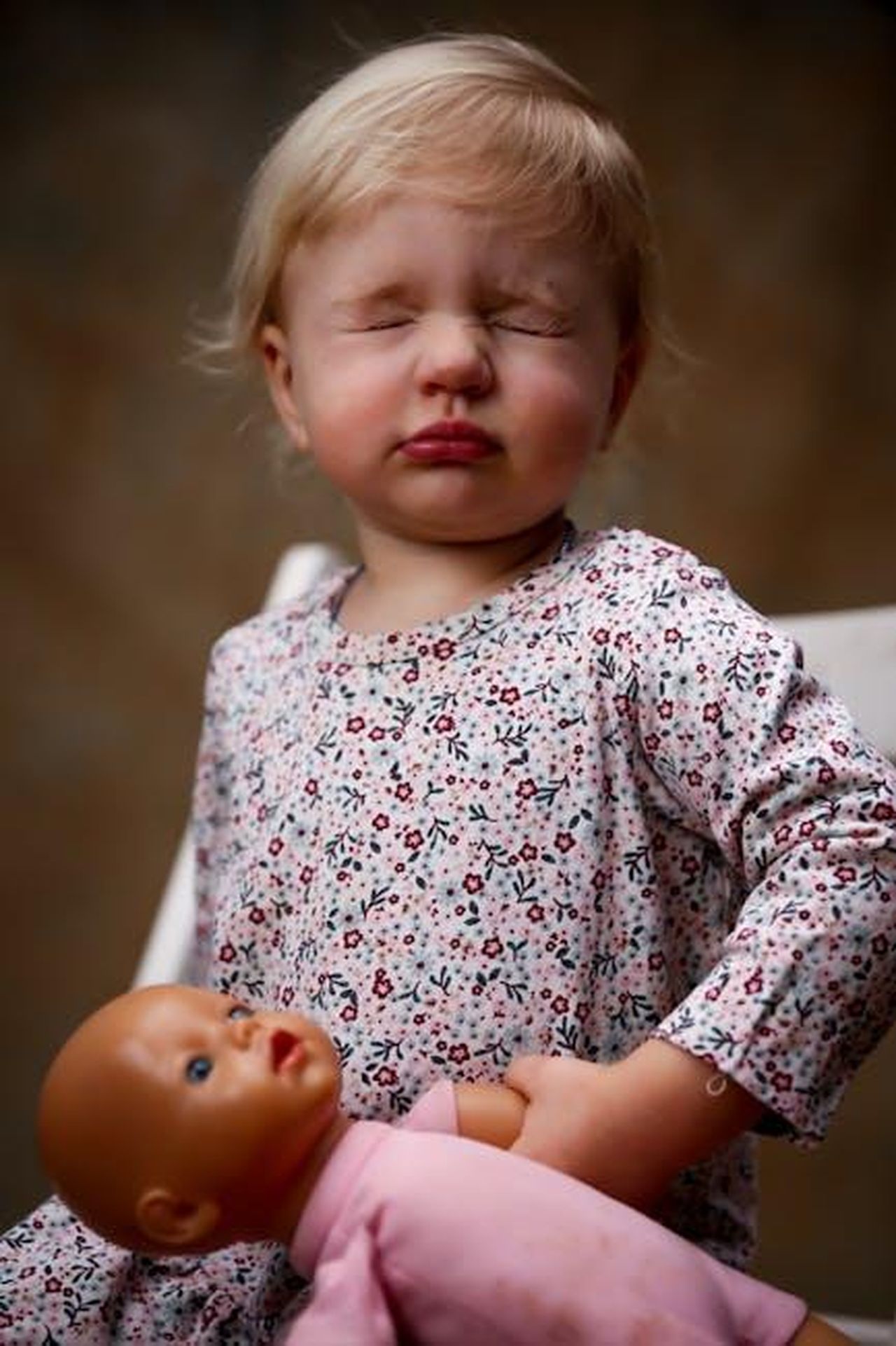 Giocando_a_nascondino_con_i_nonni_Et4NzAv5mQ - Confidenze24 Source: Саша Лазарев - pexels - Cute toddler with eyes closed, holding a doll, wearing a floral dress indoors.