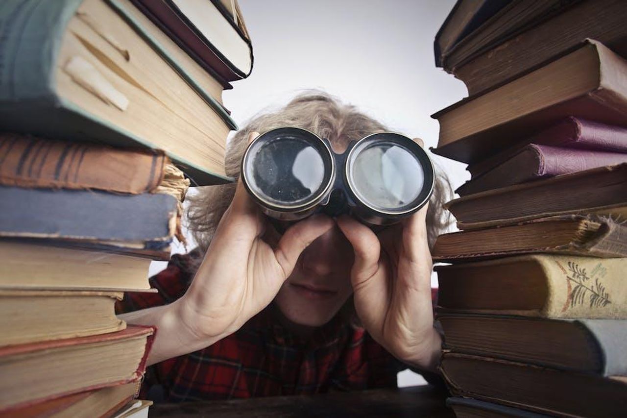Harry_Hollinger_vuole_chiudere_lindagine_troppo_in_fretta_x4K5mXJUNy - Confidenze24 Source: Andrea Piacquadio - pexels - A man observing through binoculars surrounded by stacks of vintage books indoors.