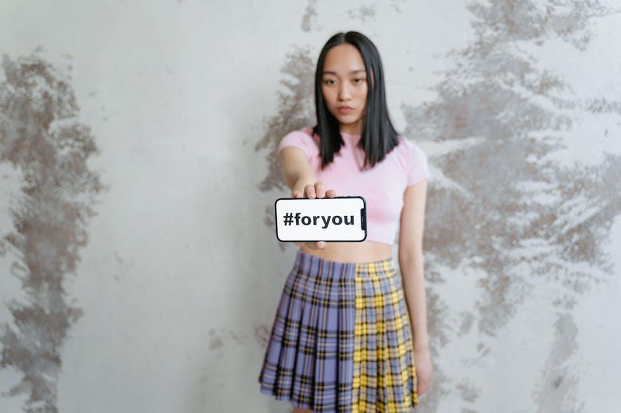 I_follower_si_uniscono_alla_polemica_u9tmE0FmfE - Confidenze24 Source: MART PRODUCTION - pexels - A young woman in a trendy outfit holds a phone displaying #foryou against a textured wall.