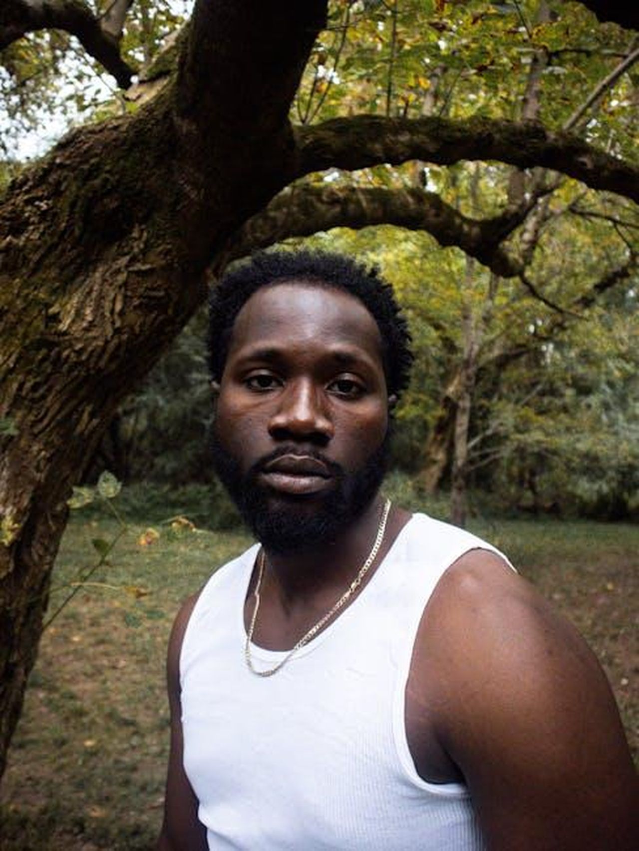 Il_cast_del_documentario_CS0m8xqYXz - Confidenze24 Source: Sherman Trotz - pexels - A thoughtful portrait of a man standing in a forest, wearing a white tank top.