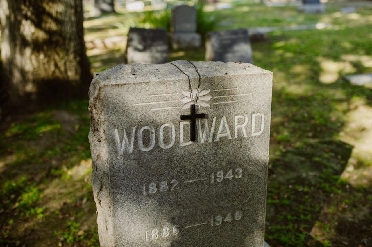 Il_cognome_Sussex_e_limportanza_della_famiglia_om1gXpmVAD - Confidenze24 Source: RDNE Stock project - pexels - A peaceful cemetery scene with a gravestone marked 'Woodward' and a cross necklace hanging in sunlight.