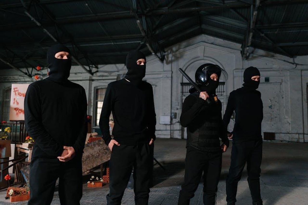 Il_contesto_della_missione_di_Billie_Matalon_70EVDyOoT5 - Confidenze24 Source: cottonbro studio - pexels - Four individuals in black attire and balaclavas standing in an industrial setting.