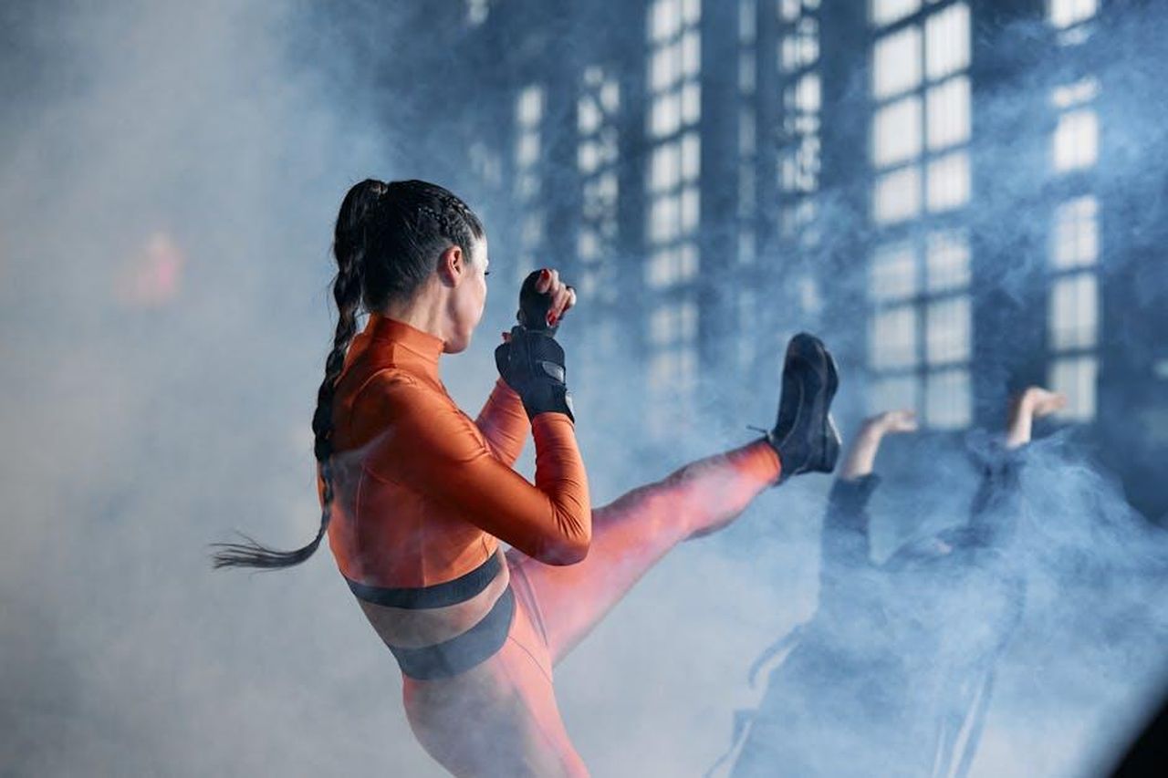 Il_contributo_di_Gene_Kelly_alla_moda_del_film_vB6N8R87RM - Confidenze24 Source: cottonbro studio - pexels - A woman in a vibrant costume performs a martial arts kick amidst smoke in an indoor setting.