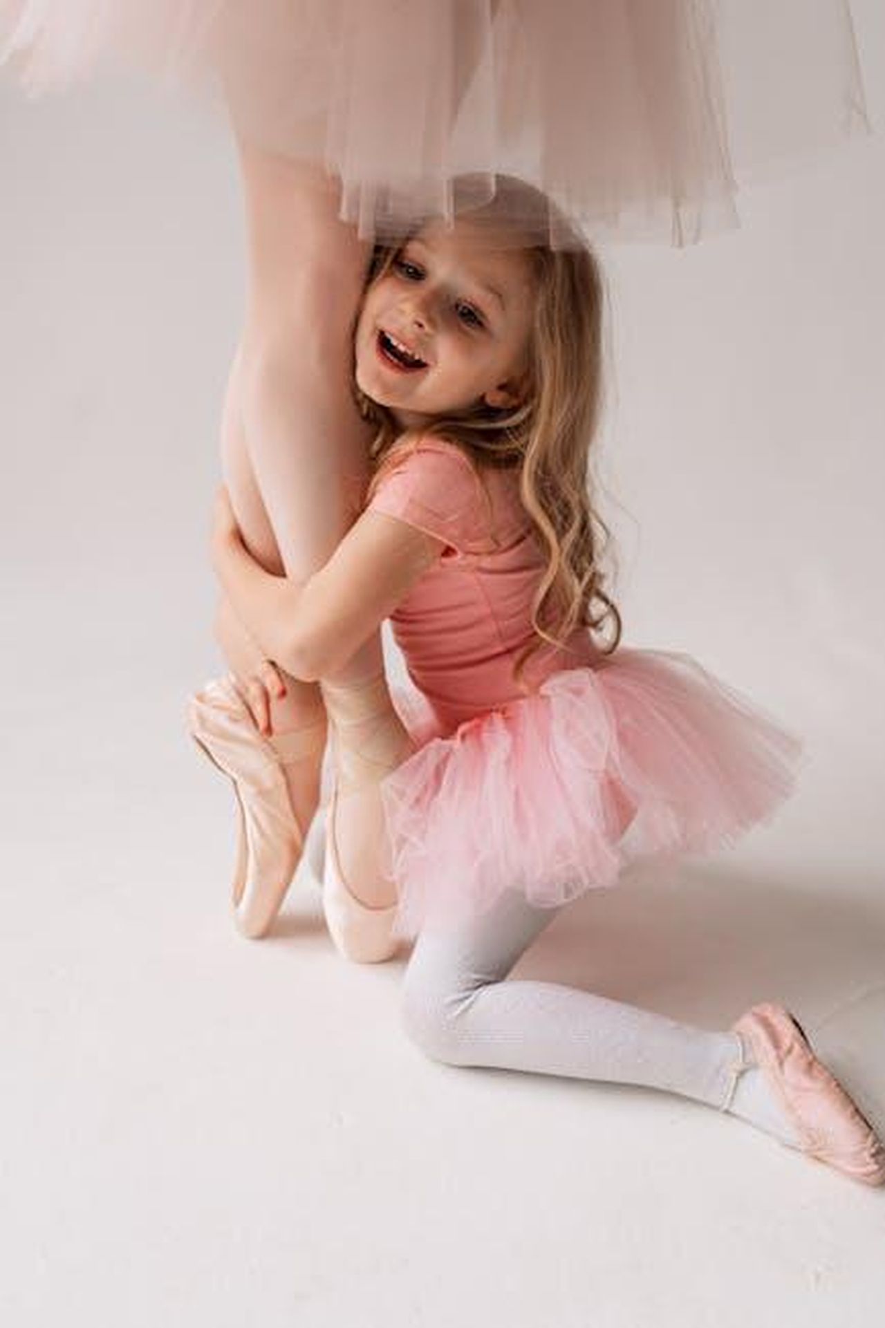 Il_legame_tra_madre_e_figlia_4yMs9oko4B - Confidenze24 Source: Victoria Strelka_ph - pexels - A smiling young girl hugs a ballerina's leg, wearing matching pink tutus in a bright studio setting.