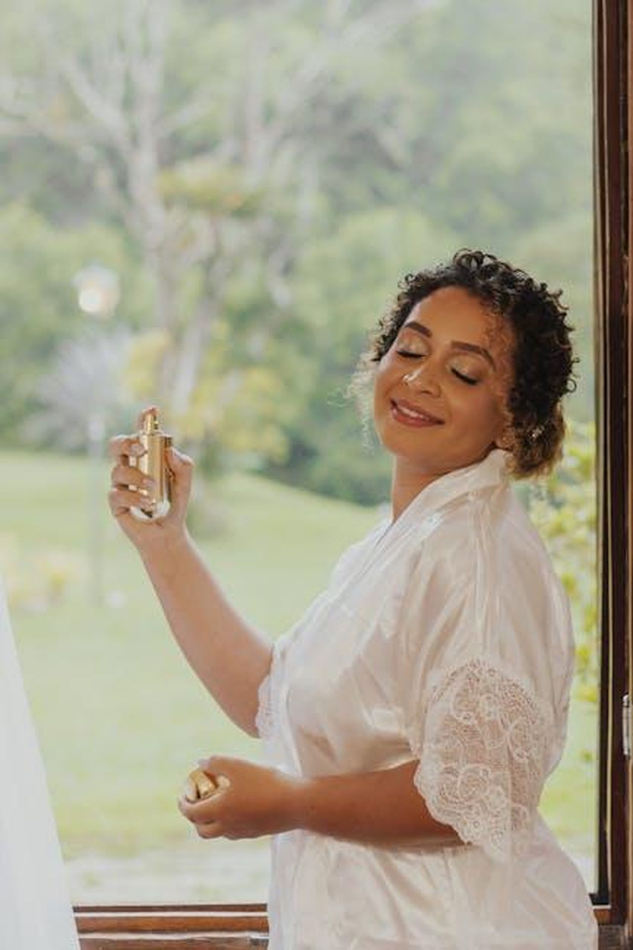 Il_messaggio_di_Keke_sulla_maternit_Xe4m2guBl0 - Confidenze24 Source: Adenir Figueiredo Carvalho - pexels - Woman in white silk robe sprays perfume, enjoying the fresh scent by a window in a serene setting.