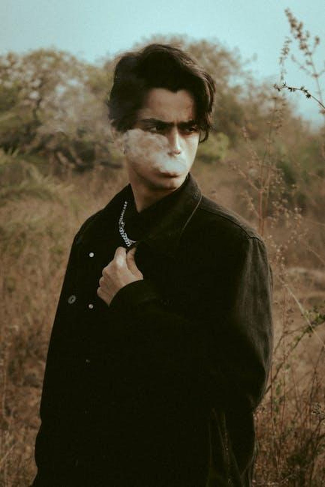 Il_mito_della_mascolinit_tossica_PhKfEBANT6 - Confidenze24 Source: Keith Lobo - pexels - A young man with dark hair smoking outdoors, exhaling smoke in a natural setting.