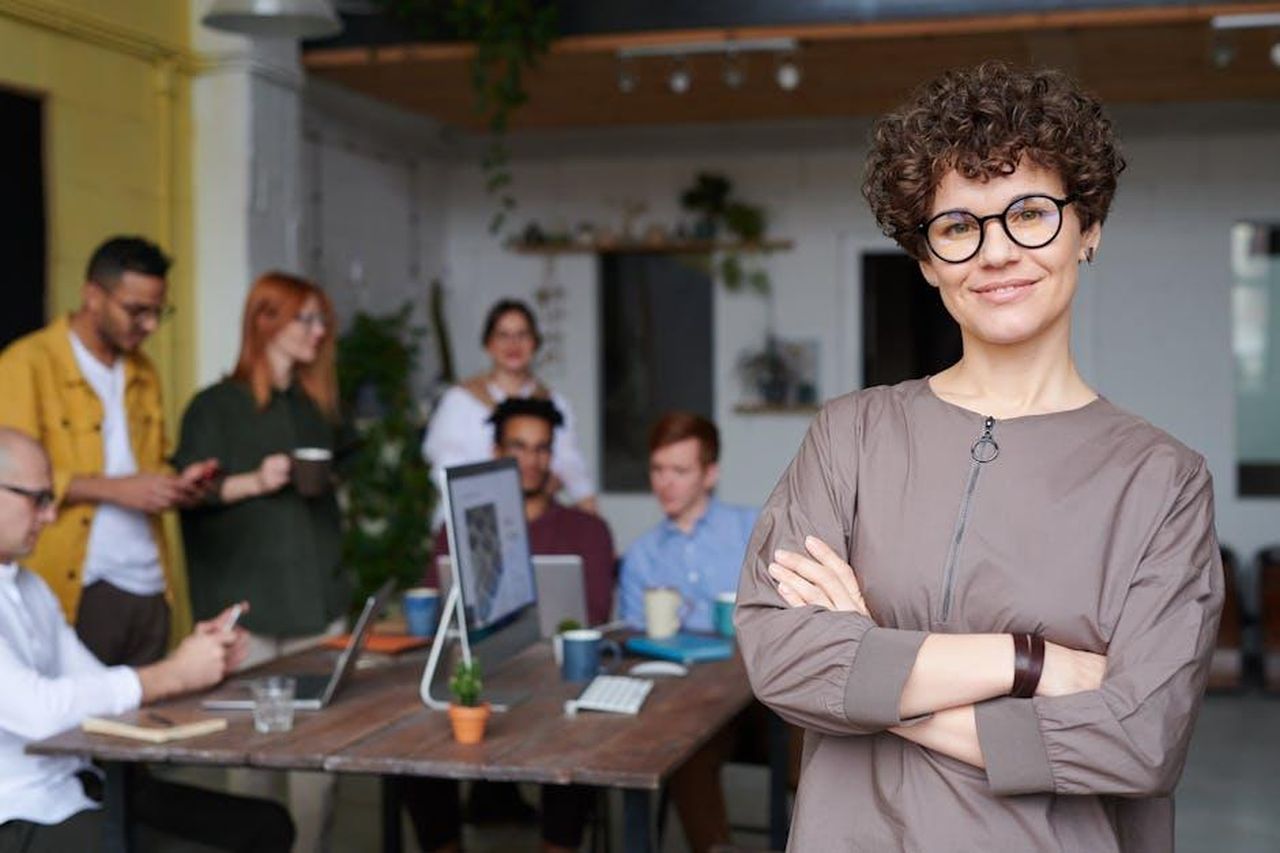 Il_ritorno_di_Foreman_e_il_suo_impero_imprenditoriale_5zvGQoLS2O - Confidenze24 Source: fauxels - pexels - Smiling businesswoman with curly hair stands confidently in a modern office space with colleagues.
