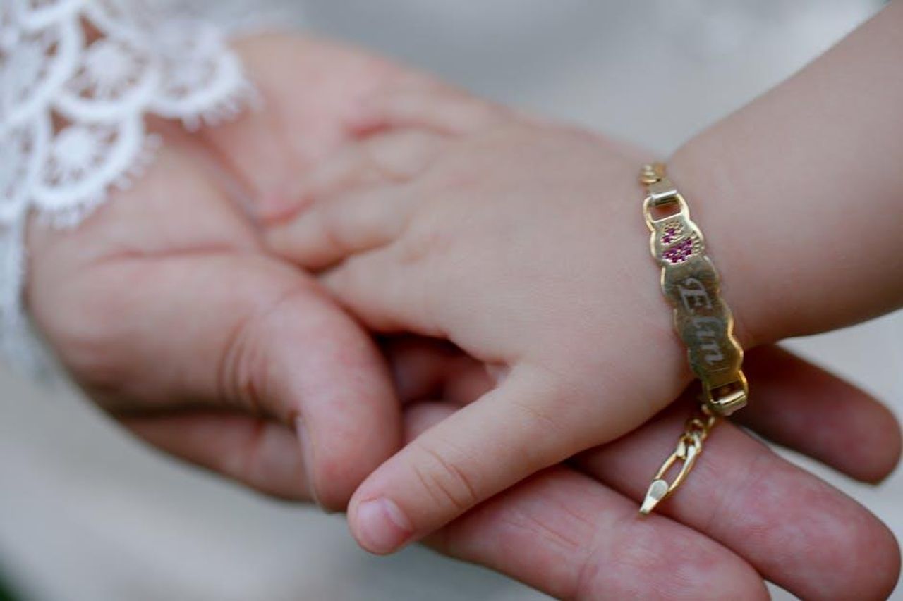 Il_significato_del_nome_Palma_D9KAWC5Odi - Confidenze24 Source: Khabbab Abdelmaqsoud - pexels - Close-up of a baby holding an adult's hand, featuring a gold bracelet with engraved name.