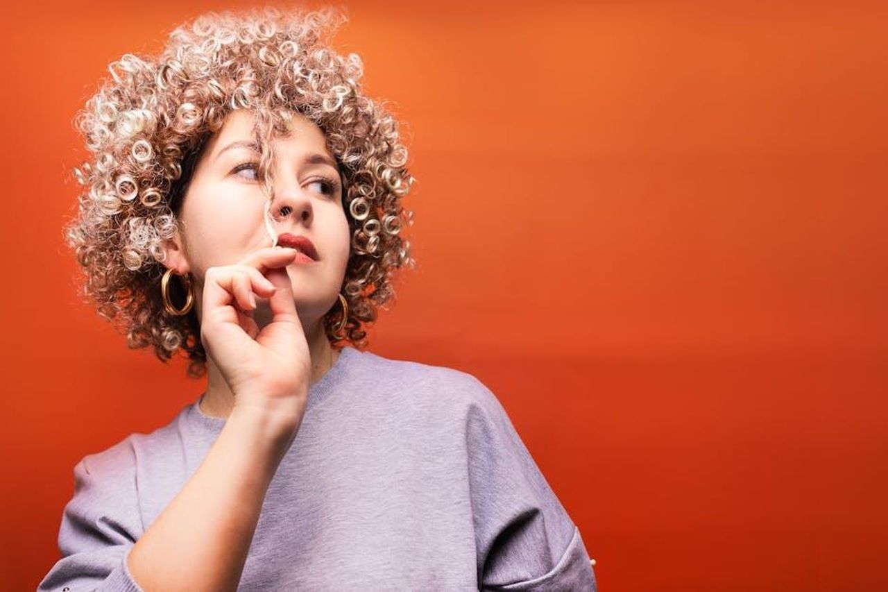Il_tono_comico_di_The_Residence_R6mG1z61lo - Confidenze24 Source: Alex P - pexels - Portrait of a young woman with curly hair, wearing a gray top, set against a vibrant red background.