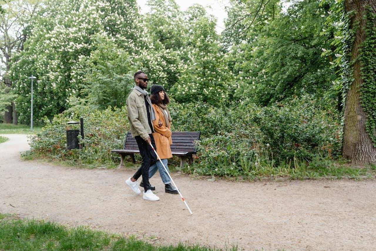 Il_viaggio_di_Taylor_e_Daniel_nella_stagione_8_YBjExtkuG5 - Confidenze24 Source: MART PRODUCTION - pexels - A visually impaired couple walks hand-in-hand with a cane through a verdant park path.