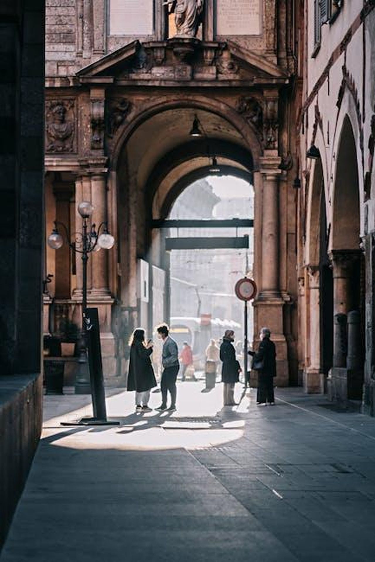 Incontro_a_Milano_e_commenti_su_Titanic_lMBTqu1L7o - Confidenze24 Source: Lorenzo Alessio Messina - pexels - People standing and walking in a sunlit historic alley under an ornate archway.