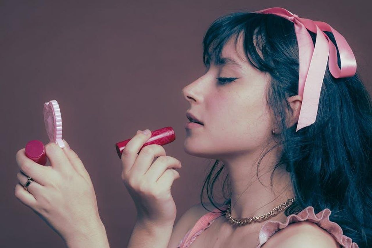 Ingredienti_chiave_e_benefici_AOCxie7Ep6 - Confidenze24 Source: Becca Correia - pexels - Portrait of a young woman applying lipstick, wearing a pink bow.