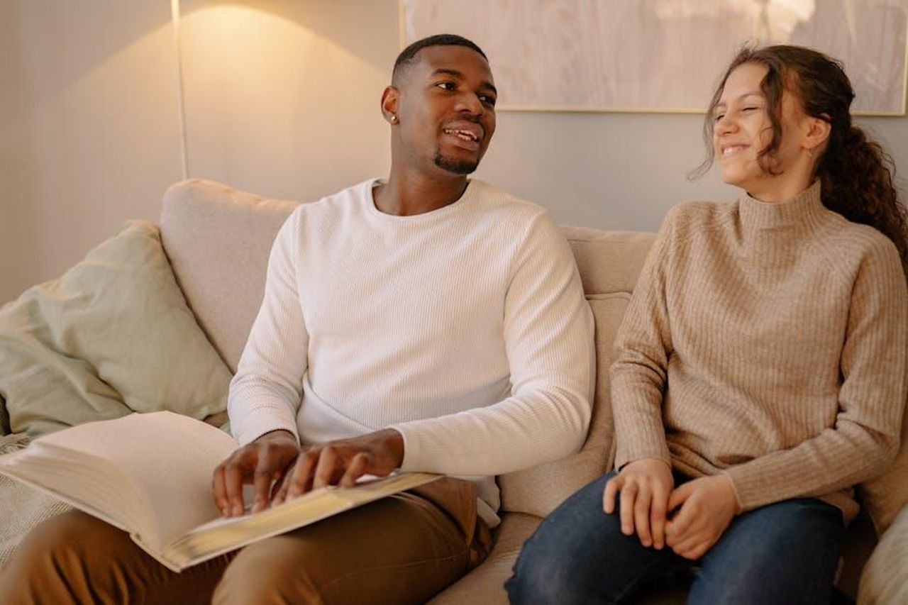 Inizio_difficile_per_Sara_e_Ben_H8m1ZQCeYM - Confidenze24 Source: Yan Krukau - pexels - A diverse couple sits on a sofa, enjoying a Braille book together in a cozy home setting.