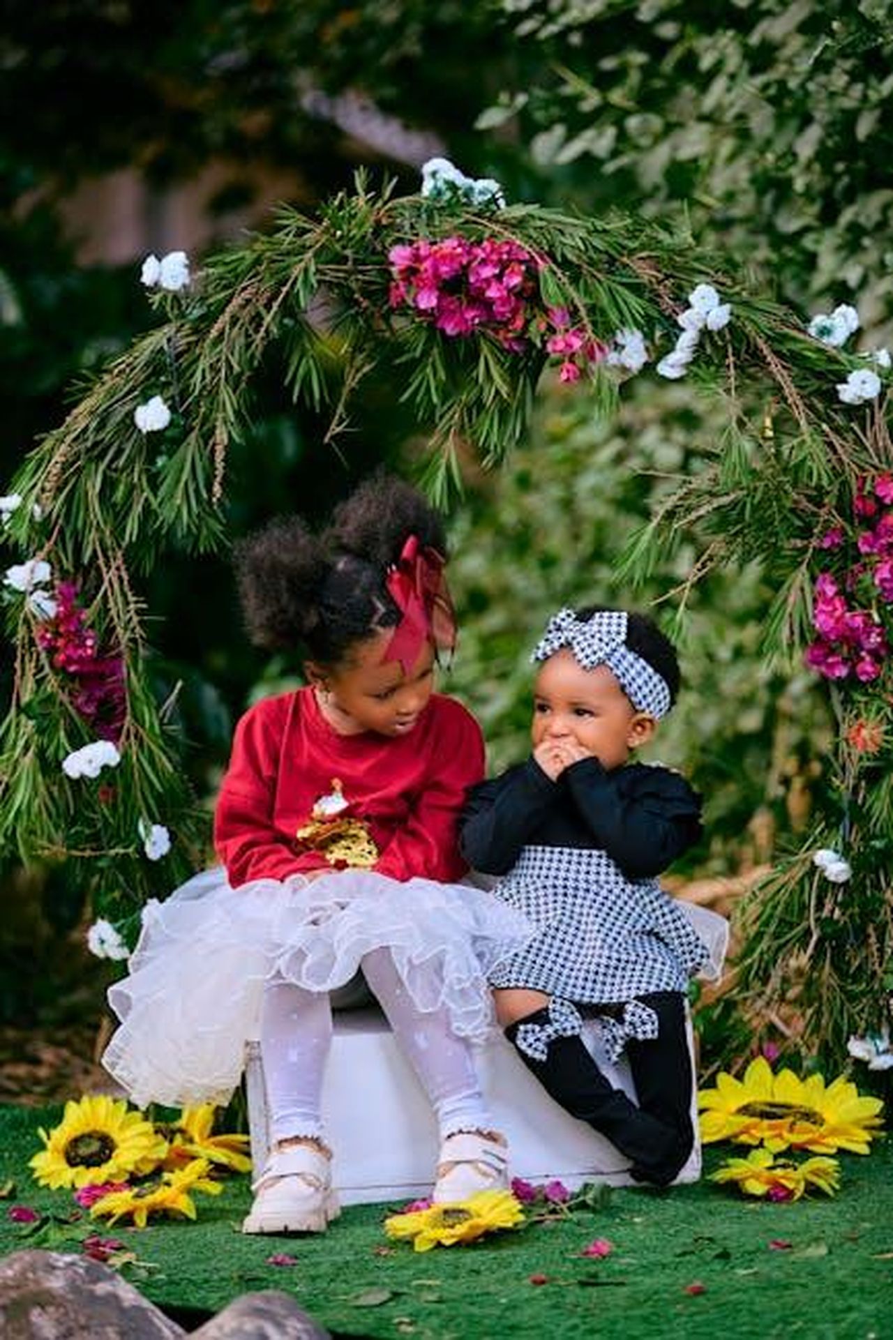 Jenna_e_la_Famiglia_gRcP2tG2Qw - Confidenze24 Source: Mika Photogenius - pexels - Two young children dressed for a festive occasion in a lush, flower-filled outdoor setting.