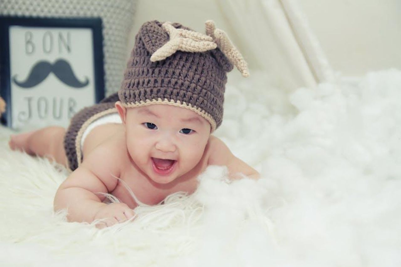 LAnnuncio_della_Nascita_di_Tove_Jane_McDowell_suHDcujh0i - Confidenze24 Source: The Craft Wonder - pexels - A joyful baby wearing a brown knitted hat lies on a soft surface, radiating happiness.
