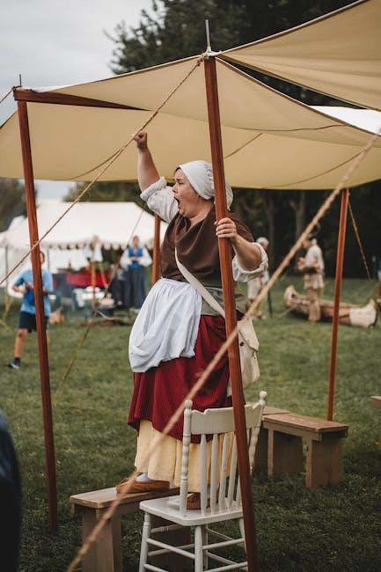 LAutenticit_del_Ruolo_di_AB_Wynter_7pKtiJ9Z7i - Confidenze24 Source: Clément Proust - pexels - A woman in period costume enthusiastically performs at an outdoor historical reenactment event under a tent.