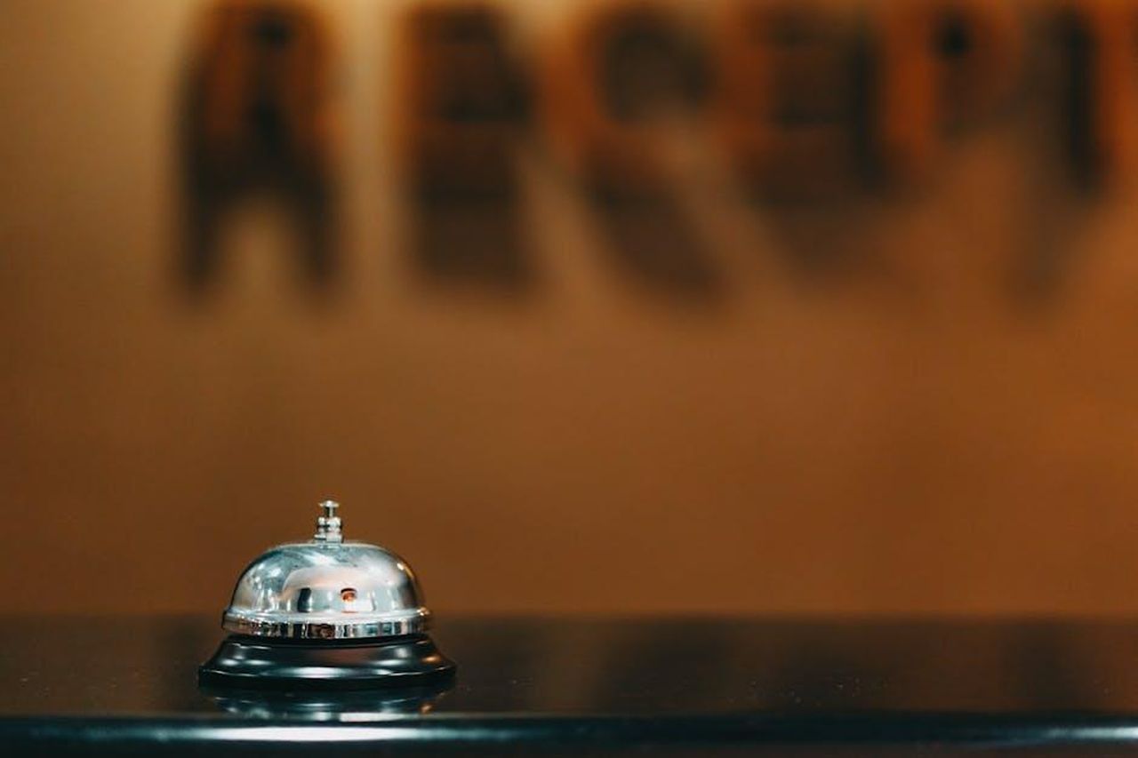 La_Reazione_dei_Fan_e_il_Passato_di_Whoopi_come_Presentatrice_mWDI666M9i - Confidenze24 Source: Mikhail Nilov - pexels - Close-up image of a silver hotel bell on a black surface with 'RECEPTION' blurred in the background.