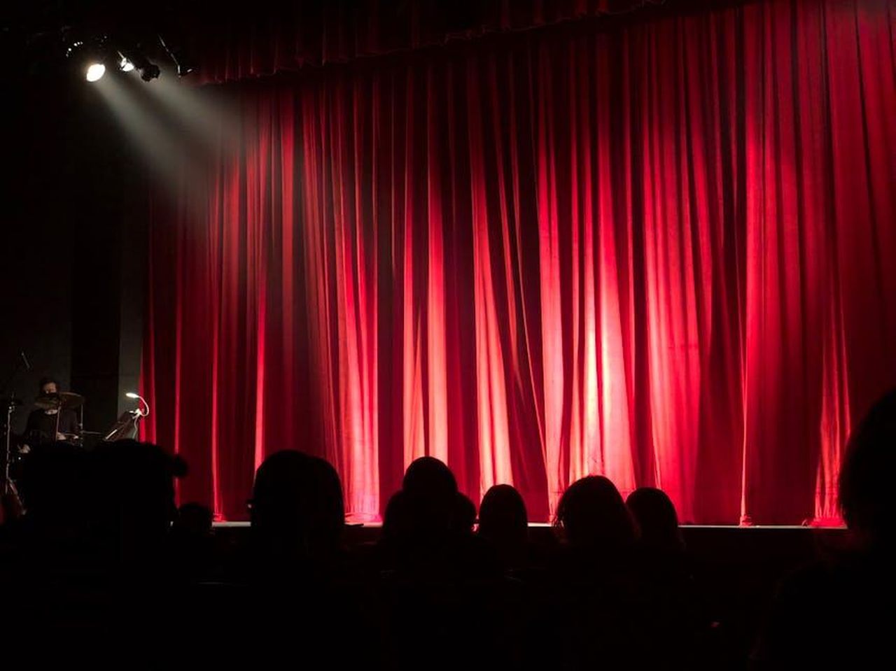 La_Reazione_di_Jenna_e_Willie_Geist_uxaffA655a - Confidenze24 Source: Monica Silvestre - pexels - Dimly lit theater stage with red curtains and audience silhouettes under spotlights.